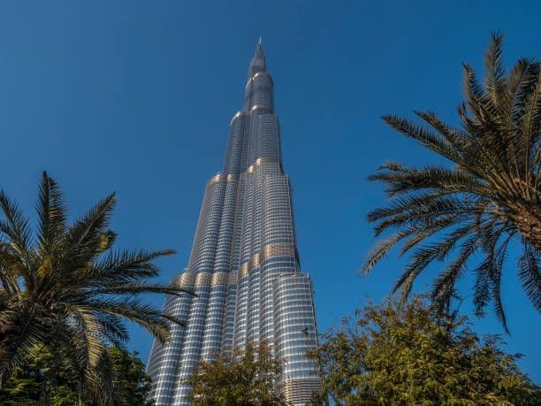 Eine Untersicht auf einen hoch aufragenden modernen Wolkenkratzer mit einer Metallfassade vor einem strahlend blauen Himmel, eingerahmt von grünen Palmen.
