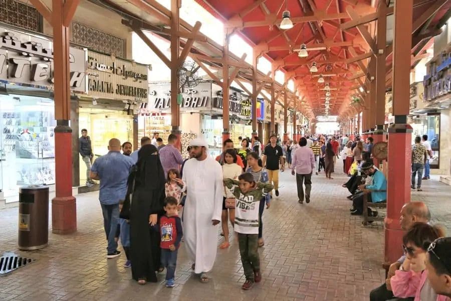 Eine belebte Menschenmenge, darunter Familien, schlendert durch einen geschäftigen, überdachten Marktplatz mit einem hohen Holzbalkendach.