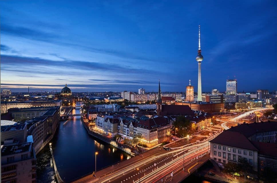 Eine Stadtlandschaft in der Abenddämmerung mit einem Fluss, einem hohen, beleuchteten Fernsehturm und Lichtspuren vom Verkehr auf einer Brücke.