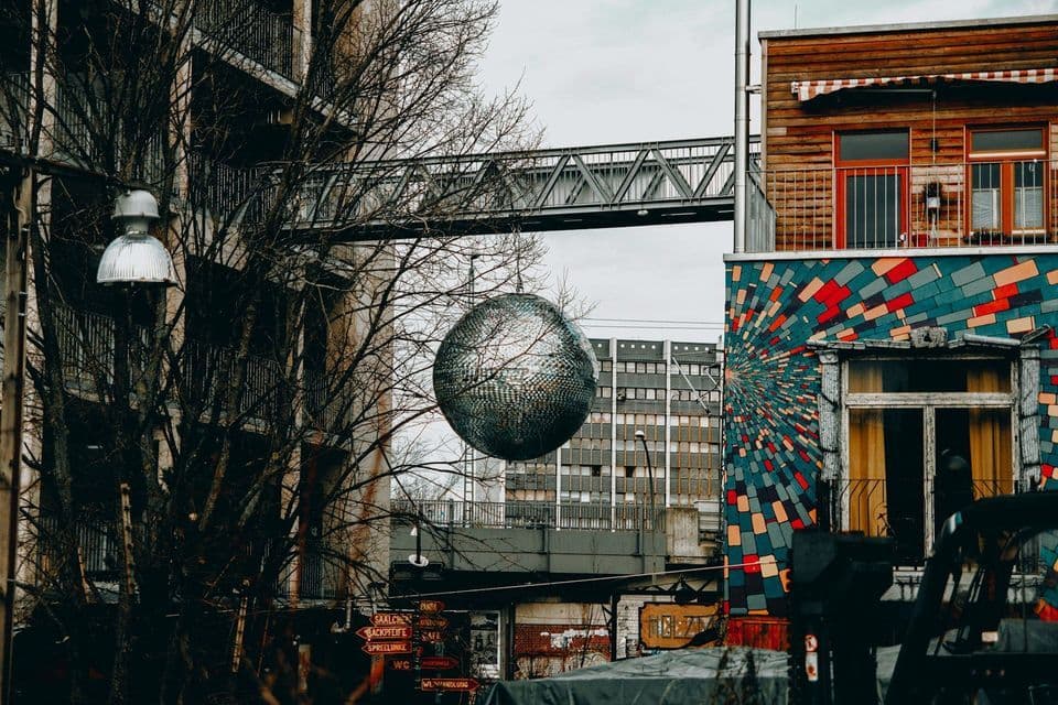 Eine große Discokugel hängt zwischen kahlen Bäumen in einem städtischen Umfeld mit einem farbenfrohen Wandbild und einem Metallsteg.