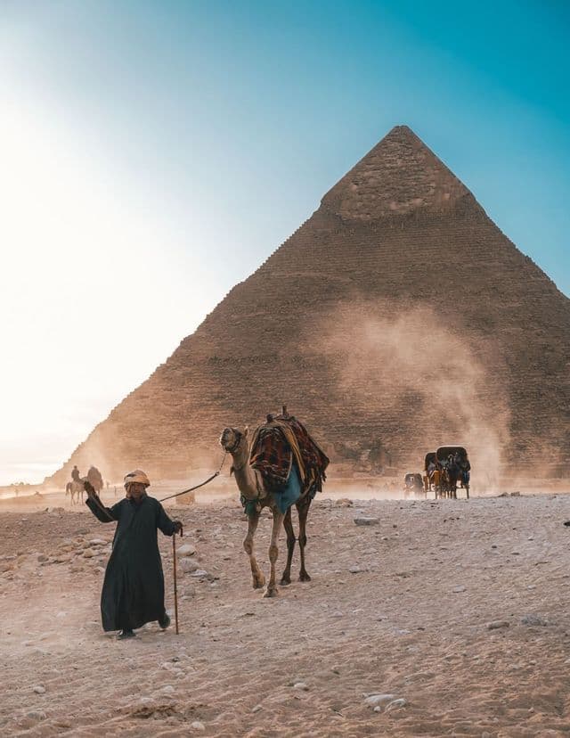 Eine Person in traditioneller Kleidung führt ein Kamel durch die Wüste, im staubigen Hintergrund ragt eine große Pyramide auf.