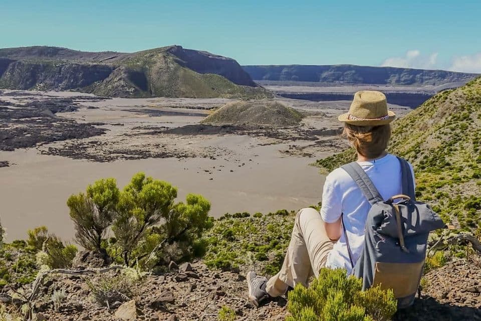 Una persona con uno zaino e un cappello siede su una collina verdeggiante, osservando un vasto paesaggio arido di crateri sotto un cielo azzurro e limpido.