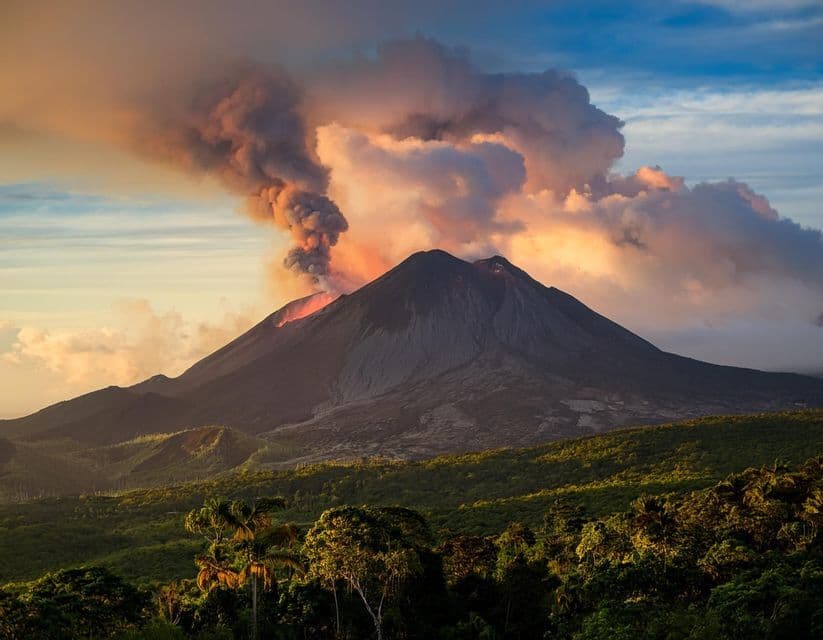 Un vulcano in eruzione emette una colonna di fumo e cenere nel cielo del tramonto, sovrastando una lussureggiante foresta verde.