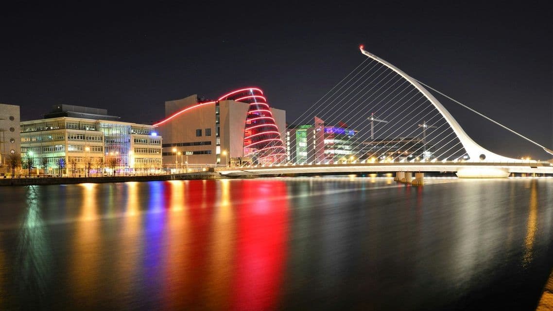 Eine moderne, beleuchtete Schrägseilbrücke überspannt nachts einen Fluss, wobei sich bunte Stadtlichter auf der Wasseroberfläche spiegeln.