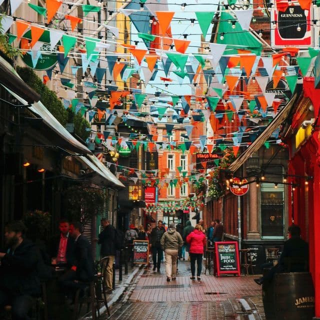 Eine schmale, belebte Stadtstraße, geschmückt mit grünen, weißen und orangefarbenen Wimpelketten, die zwischen den Gebäuden gespannt sind.