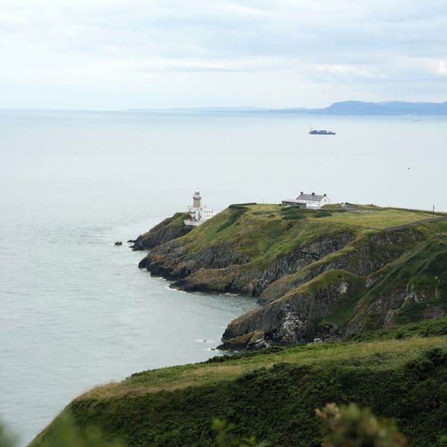 Ein weißer Leuchtturm und ein Gebäude thronen auf einer grünen, felsigen Klippe mit Blick auf ein ruhiges Meer unter einem bewölkten Himmel.