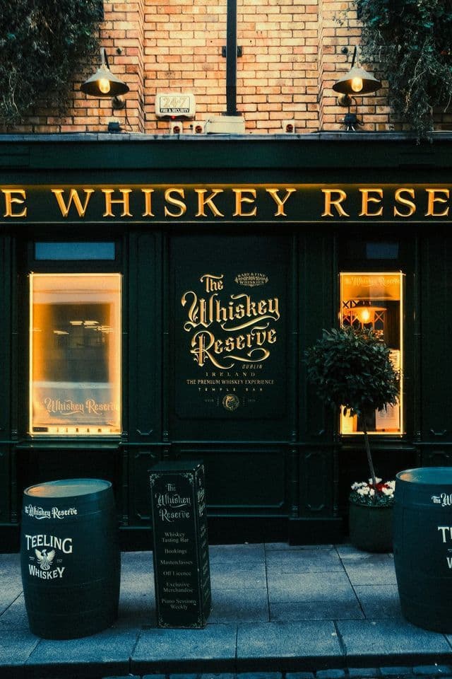 Die dunkelgrüne Fassade der Bar 'The Whiskey Reserve', beleuchtet von warmen Lichtern, mit Whiskeyfässern und einem Schild auf dem Gehweg.
