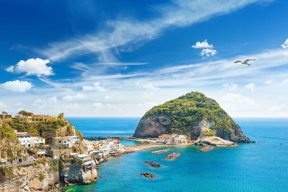 A coastal village on a cliff overlooks a bay with a small beach and a green, rocky island under a bright blue sky.