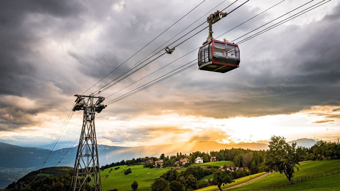 Eine Seilbahn gleitet an ihren Drahtseilen über ein grünes Bergtal mit Häusern, unter einem bewölkten Himmel bei Sonnenuntergang.