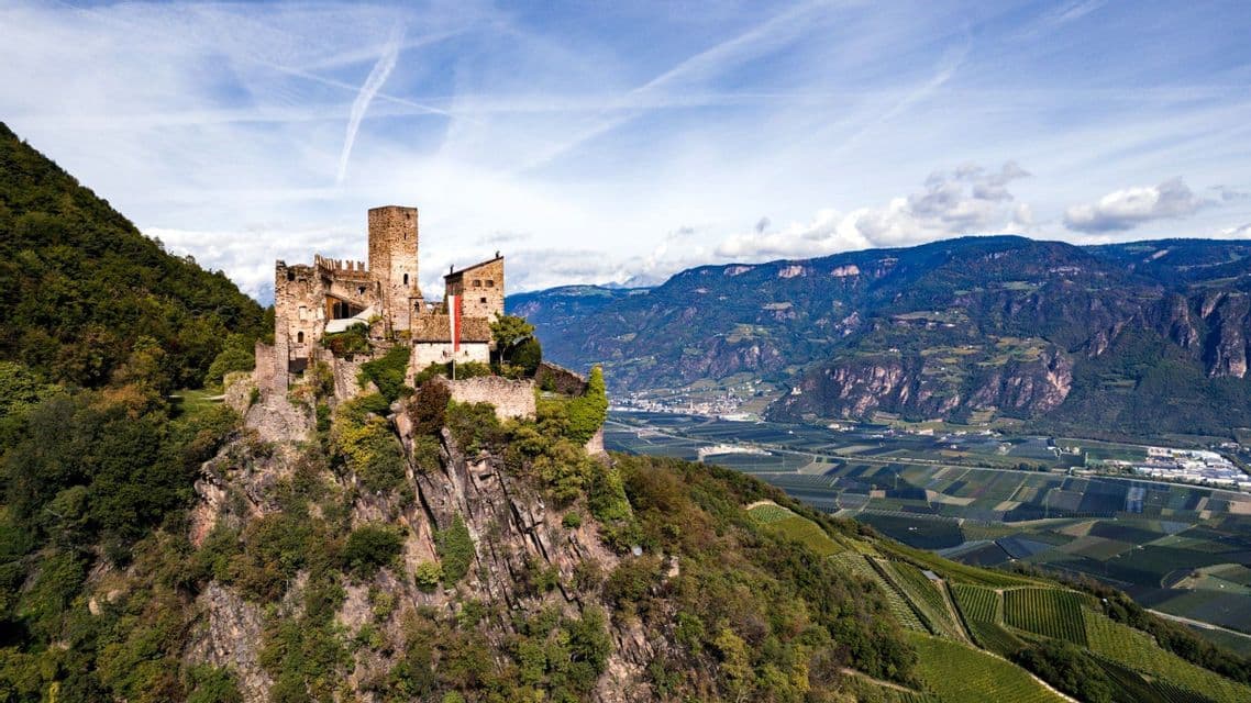 Eine Steinburg thront auf einem felsigen Kliff und überblickt ein weites Tal mit Weinbergen und Bergen in der Ferne.