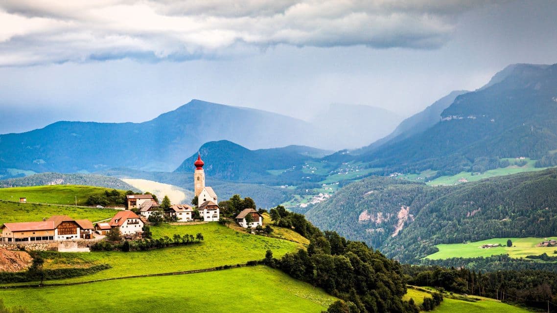 Eine Kirche mit rotem Zwiebelturm und ein kleines Dorf liegen auf einem grünen Hügel mit Blick auf ein weites Gebirgstal unter einem bewölkten Himmel.