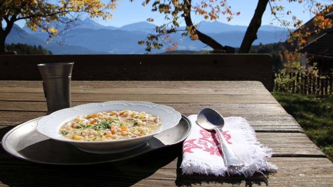 Eine Schale Gersten-Gemüse-Suppe auf einem Holztisch im Freien, mit einer Bergkette im Hintergrund unter klarem Himmel sichtbar.
