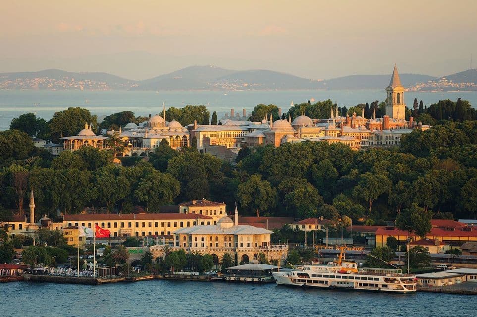 A historic palace complex with domes and towers sits on a lush, green hill overlooking the water at sunset.