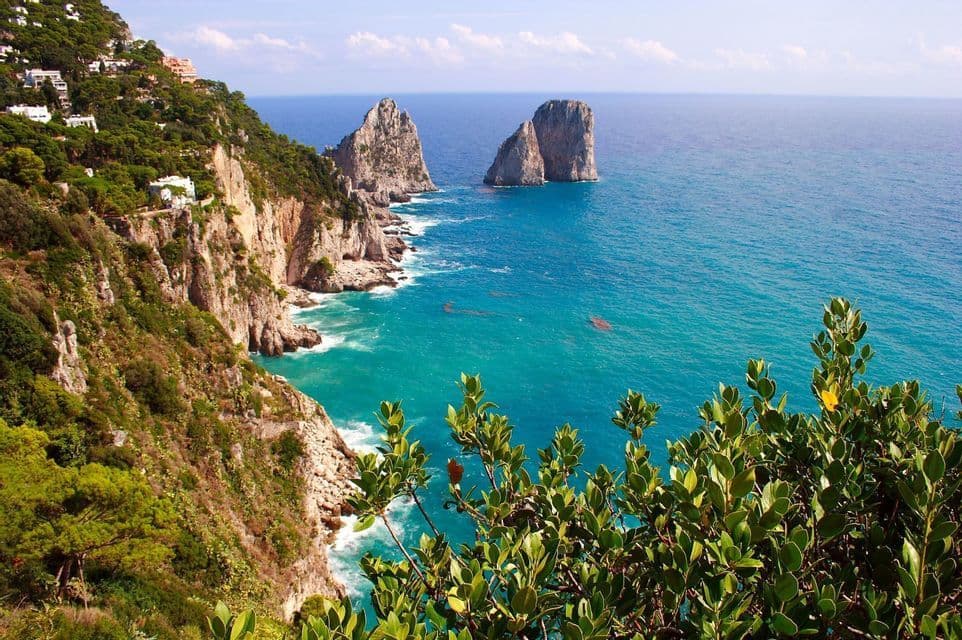 View of two large rock formations in a turquoise sea from a lush, green cliffside dotted with white buildings.