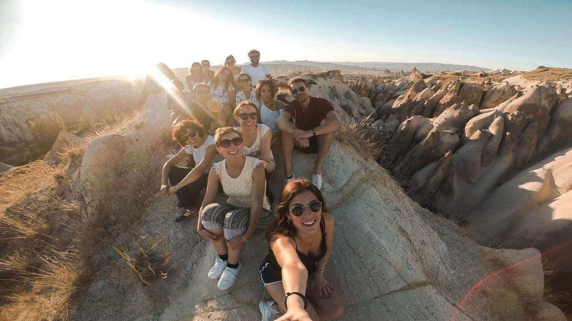 Un groupe WeRoad prend un selfie sur un pic rocheux, avec un vaste canyon de formations rocheuses en arrière-plan, au coucher du soleil.