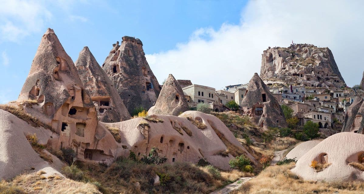 Un pueblo de casas cueva excavadas en formaciones rocosas cónicas en la ladera de una montaña, bajo un cielo parcialmente nublado.