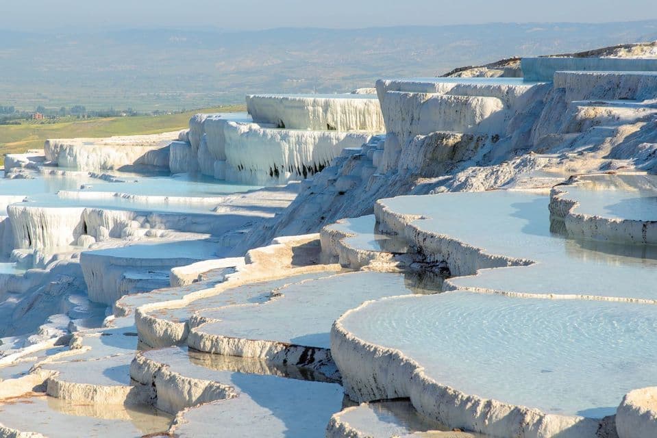 Sunlight illuminates white travertine terraces filled with pools of pale blue water on a hillside.