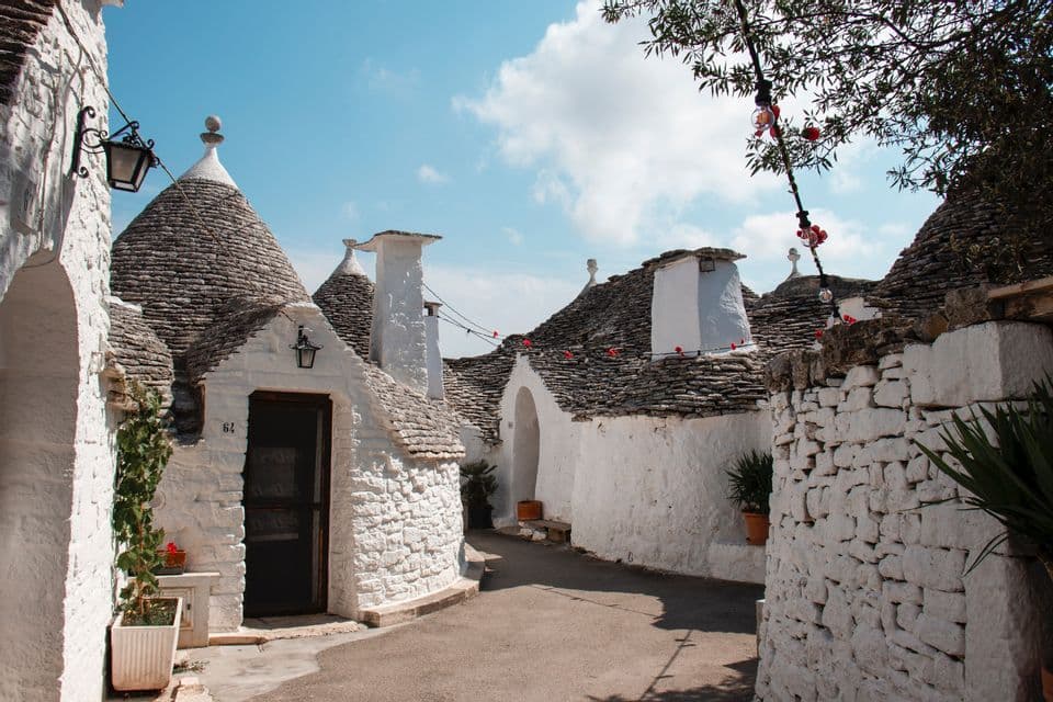 Whitewashed stone houses with conical roofs line a narrow, paved alley under a blue sky with some clouds.
