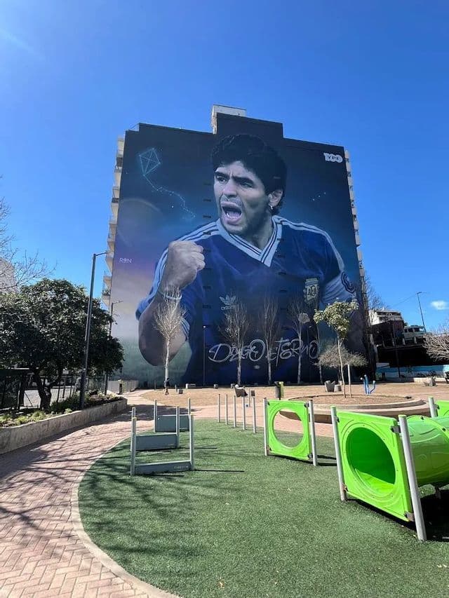 Un grande murale del calciatore Diego Maradona che festeggia su un palazzo, visto da un piccolo parco giochi.