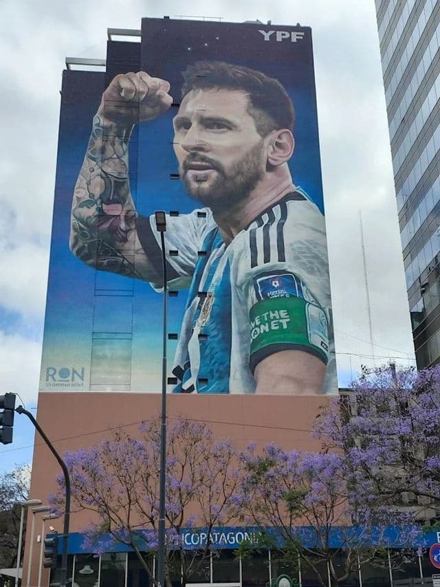 Un gigantesco murale del calciatore Lionel Messi con la maglia dell'Argentina e un pugno alzato, dipinto sul fianco di un alto edificio.