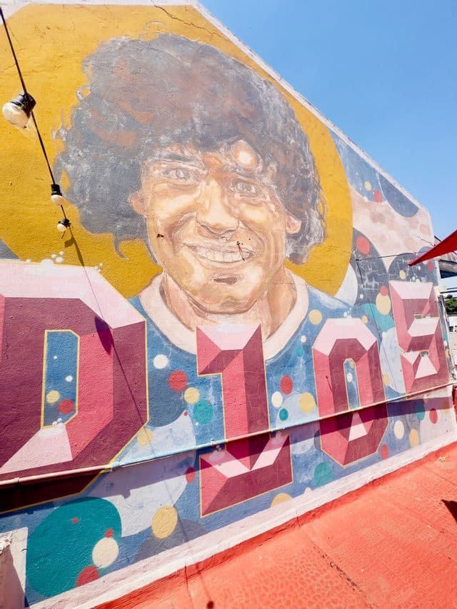 Un murale colorato di Diego Maradona è dipinto su una parete esterna, con la parola stilizzata 'D10S' a grandi lettere sotto il suo ritratto.