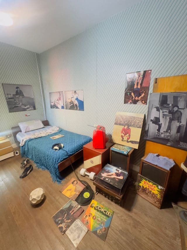 Una camera da letto in stile retrò, arredata con mobili vintage, poster sulle pareti a righe e dischi in vinile sul pavimento e sugli altoparlanti.