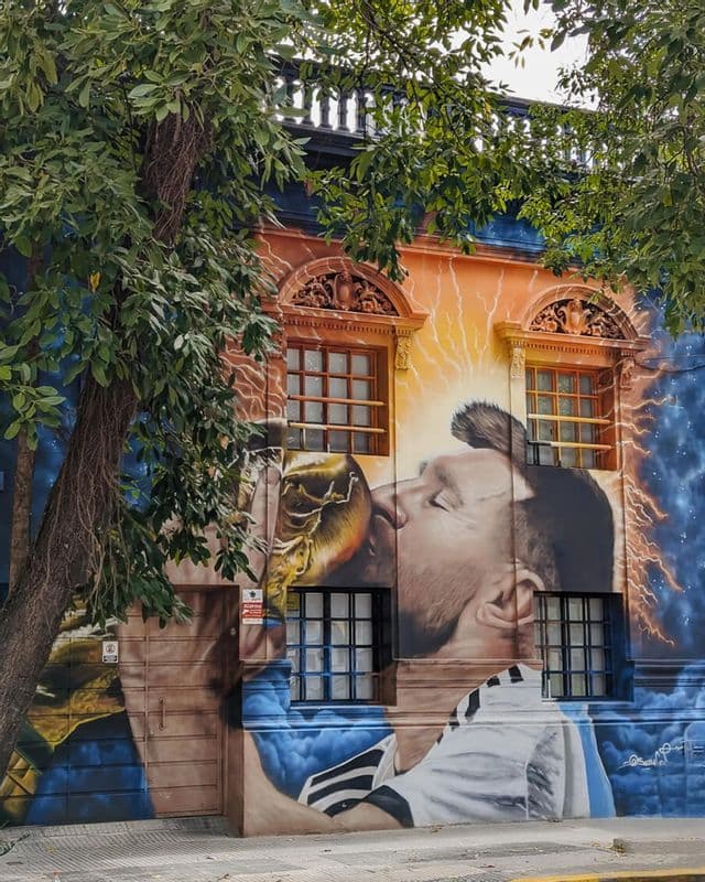 Un grande murale sul lato di un edificio raffigura un calciatore con una maglia a strisce che bacia un trofeo d'oro.