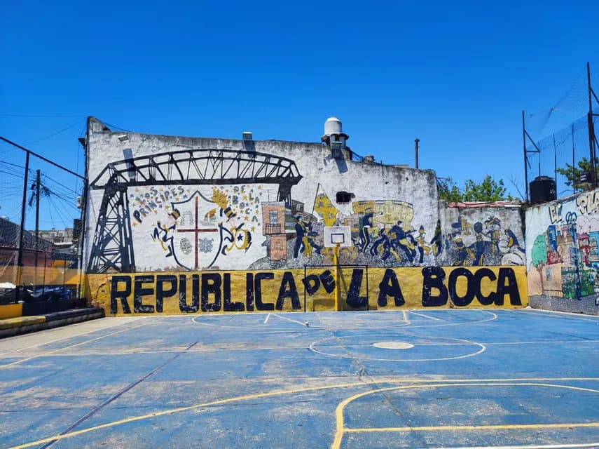 Un campo da basket blu vuoto davanti a un muro con un grande murale che recita 'Republica de La Boca' sotto un cielo sereno.