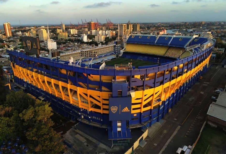 Una vista aerea di un grande stadio di calcio blu e giallo inserito in un paesaggio urbano, con un porto visibile sullo sfondo.