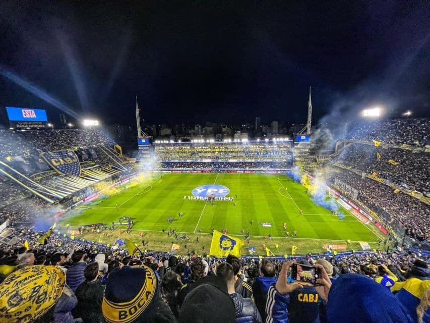 Vista grandangolare di uno stadio di calcio affollato di notte, con tifosi sugli spalti e giocatori sul campo illuminato.