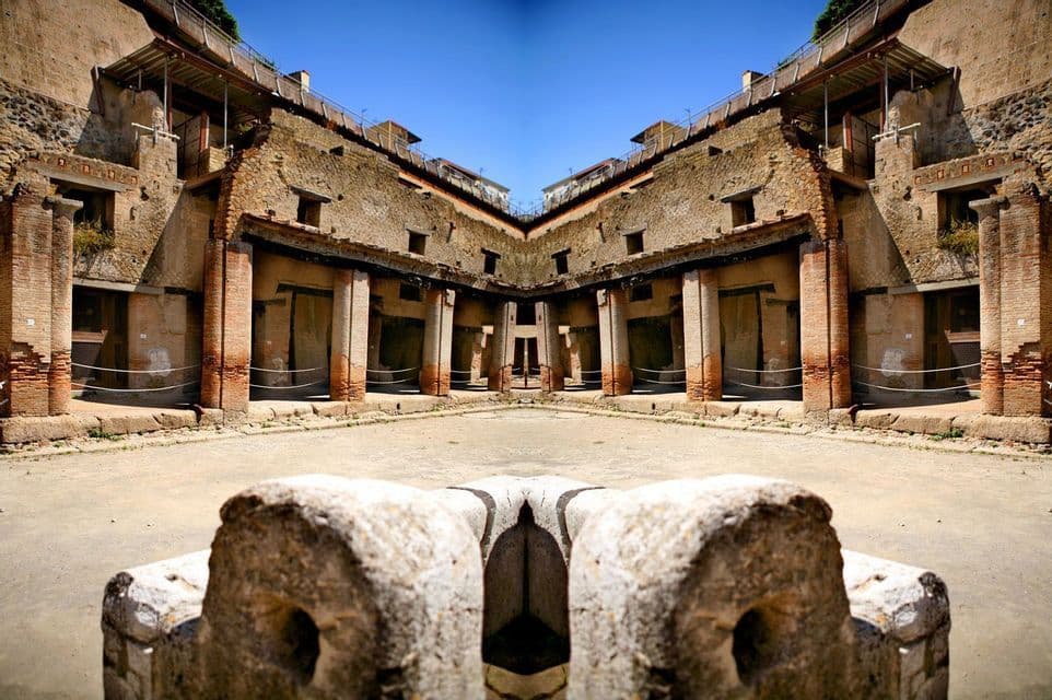 Una vista specchiata e simmetrica del cortile di un'antica rovina con pilastri in mattoni e muri in pietra sotto un cielo azzurro chiaro.
