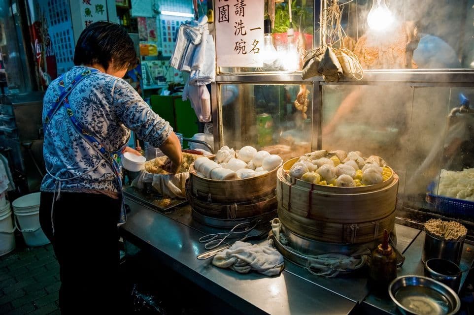 A vendor works at a night market food stall, preparing food next to two large bamboo steamers filled with steaming buns and dumplings.