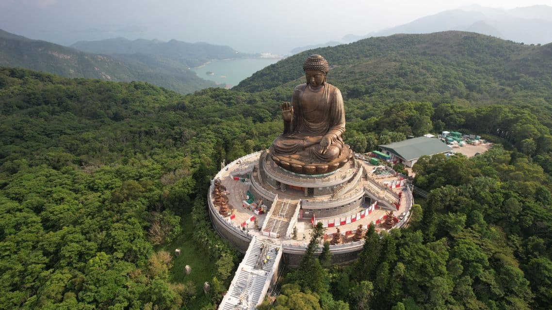 An aerial view of a giant bronze Buddha statue seated on a circular platform amidst lush green mountains and a distant lake.