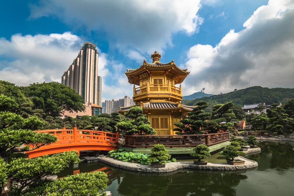 A golden pagoda on an island in a garden pond, connected by an orange bridge, with modern city skyscrapers in the background.