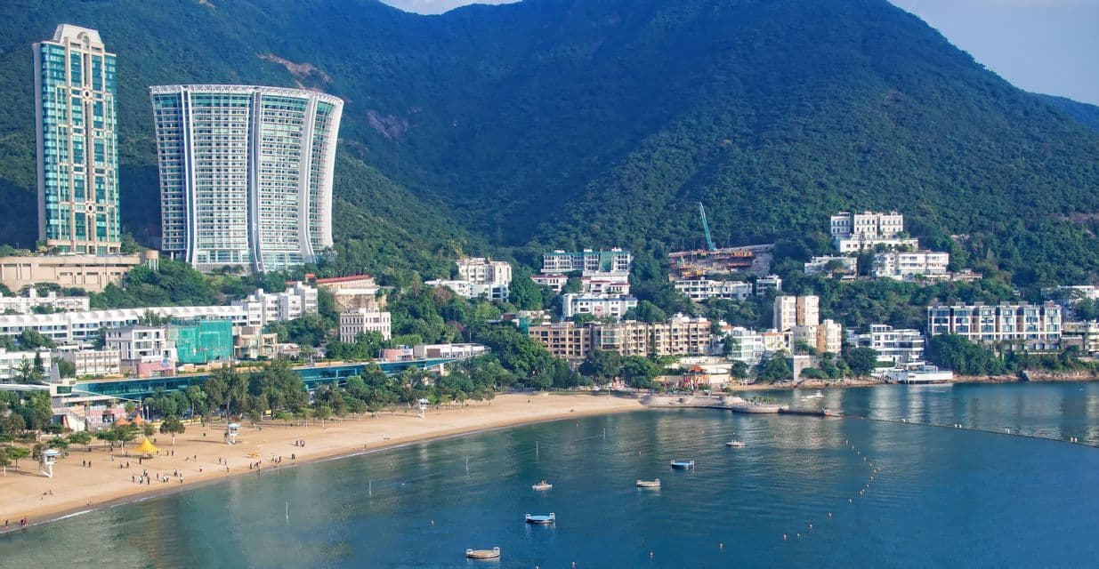 An aerial view of a coastal city with a sandy beach, modern skyscrapers, and buildings nestled into a lush green mountain.