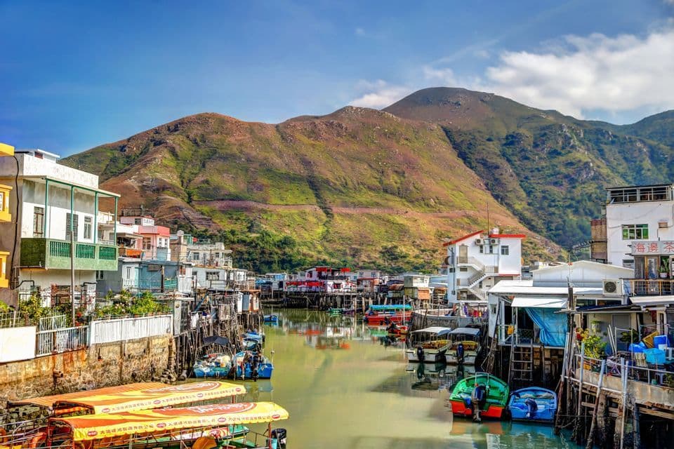 A colorful village of stilt houses and boats sits on a waterway at the base of a large, green mountain under a blue sky.