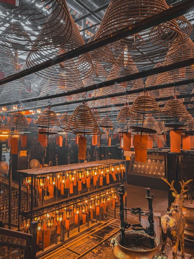 Large conical incense coils hang from the ceiling of a temple, with glowing orange lanterns and a deer statue below.
