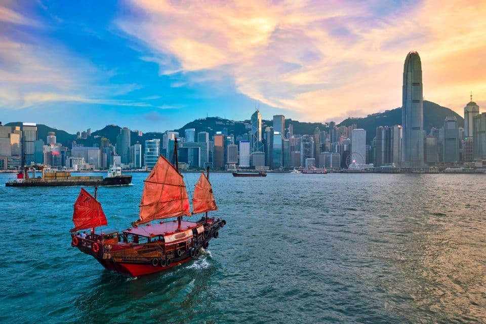 A traditional junk boat with red sails floats in a harbor with a modern city skyline in the background at sunset.