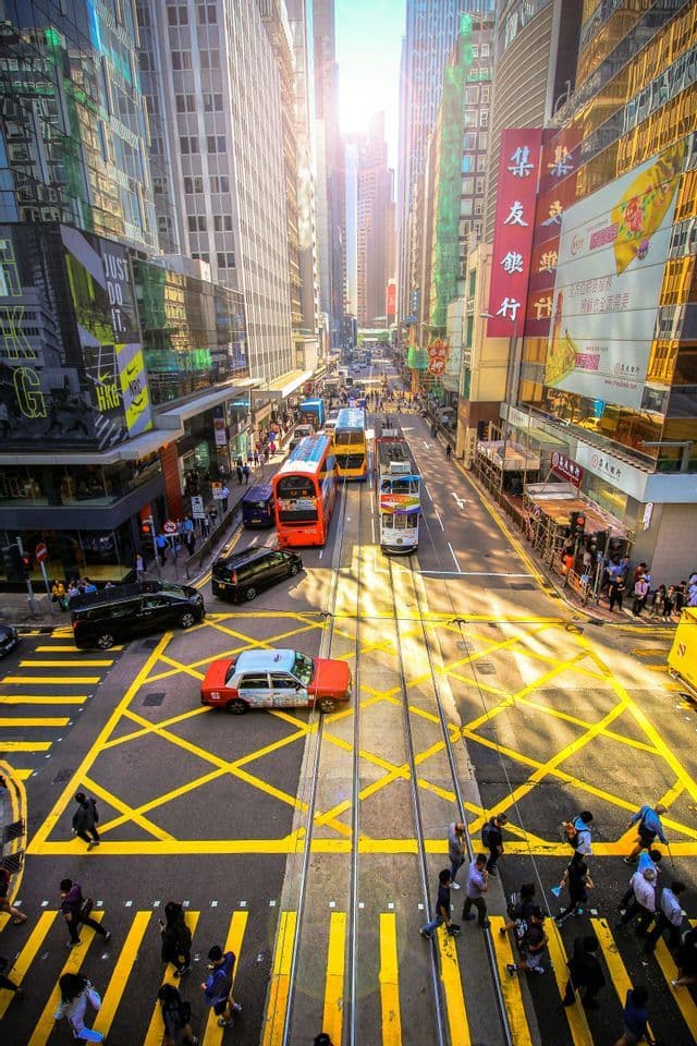A high-angle view of a bustling city street with trams, double-decker buses, and pedestrians moving between tall skyscrapers.