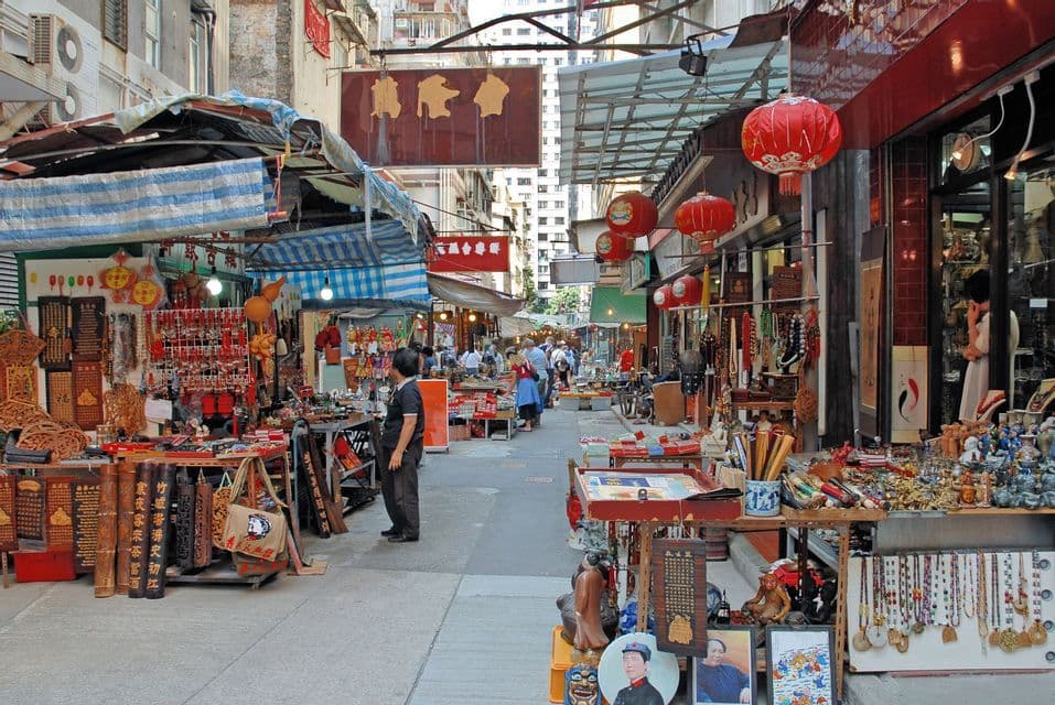 A bustling outdoor street market with stalls selling traditional crafts, souvenirs, and antiques to browsing customers under red lanterns.