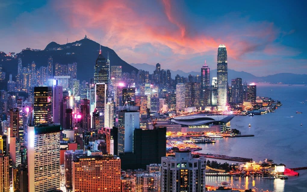 A dense city skyline with illuminated skyscrapers along a waterfront at sunset, with mountains in the background.