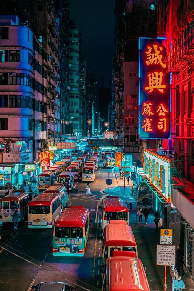 An elevated view of a busy city street at night, with traffic of red minibuses and buildings lit by colorful neon signs.