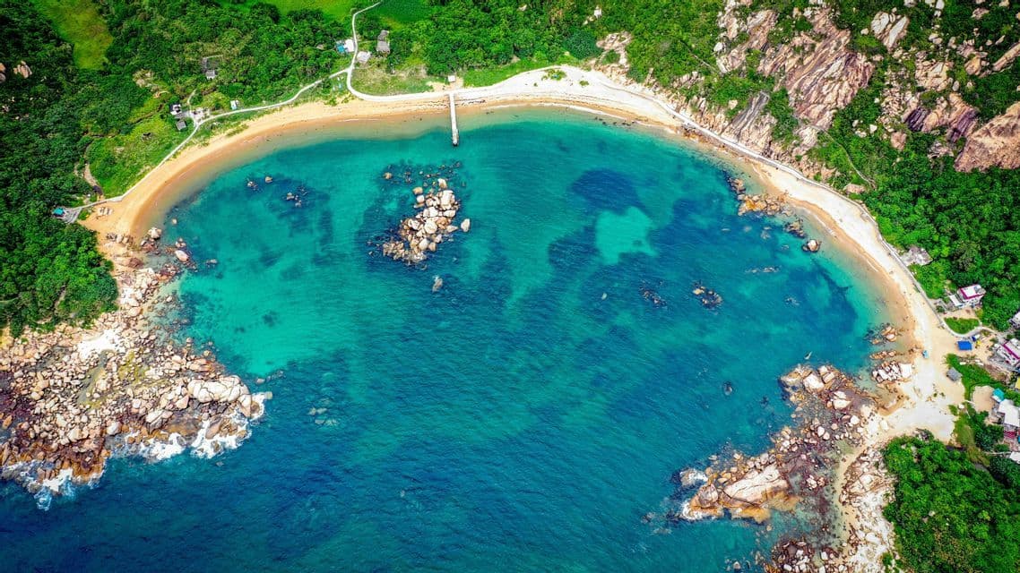 An aerial top-down view of a secluded cove with a sandy beach, clear turquoise water, and rocky, green cliffs.