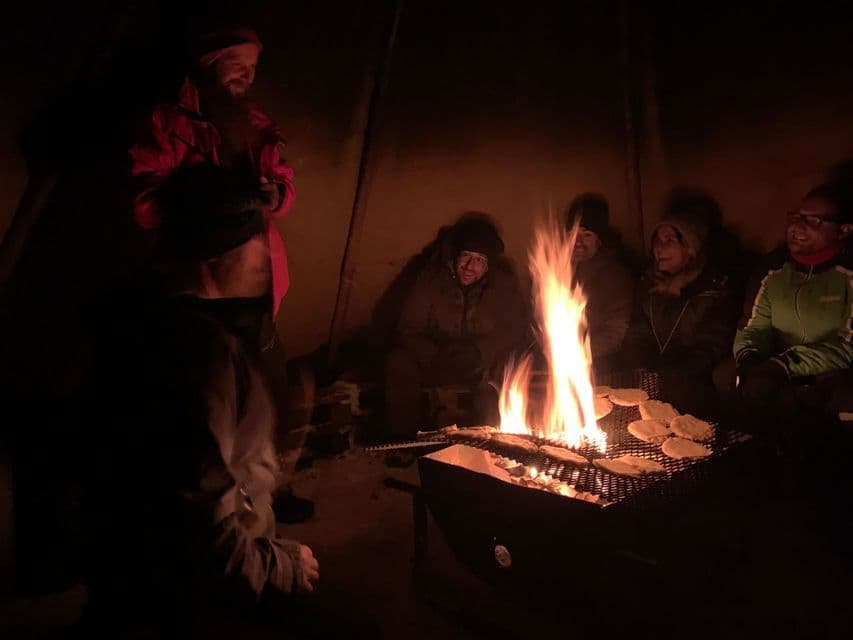 Un voyage de groupe WeRoad se rassemble autour d'un feu de camp la nuit, préparant à manger sur un grill à l'intérieur d'une tente sombre.