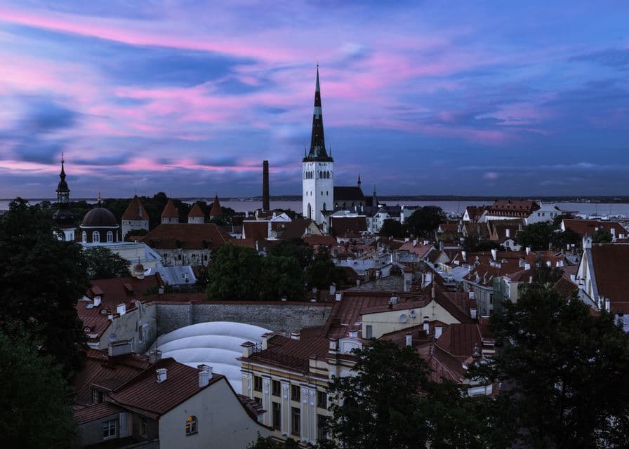 Eine europäische Stadtsilhouette mit einem markanten Kirchturm und rotgedeckten Gebäuden unter einem rosa-violetten Sonnenuntergangshimmel.