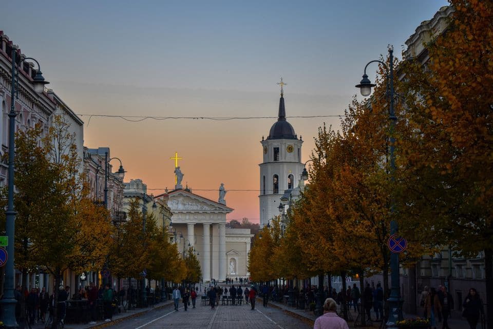 Une large rue bordée d'arbres avec un feuillage d'automne mène à une cathédrale et un clocher au crépuscule, avec des gens marchant au loin.