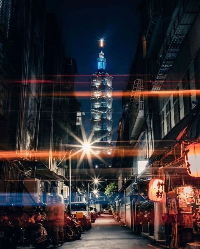 Un alto grattacielo illuminato, visto di notte da uno stretto vicolo di città, con scooter parcheggiati, lanterne luminose e scie di luce.