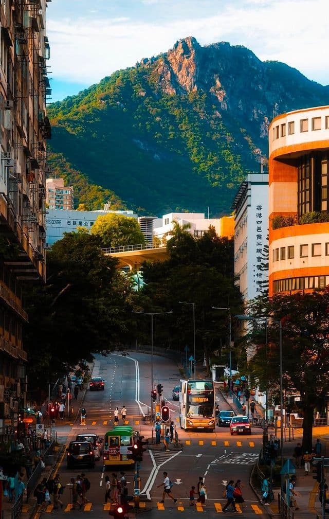 Una strada cittadina trafficata con auto, un autobus e pedoni, con una grande montagna verde che si erge sullo sfondo.