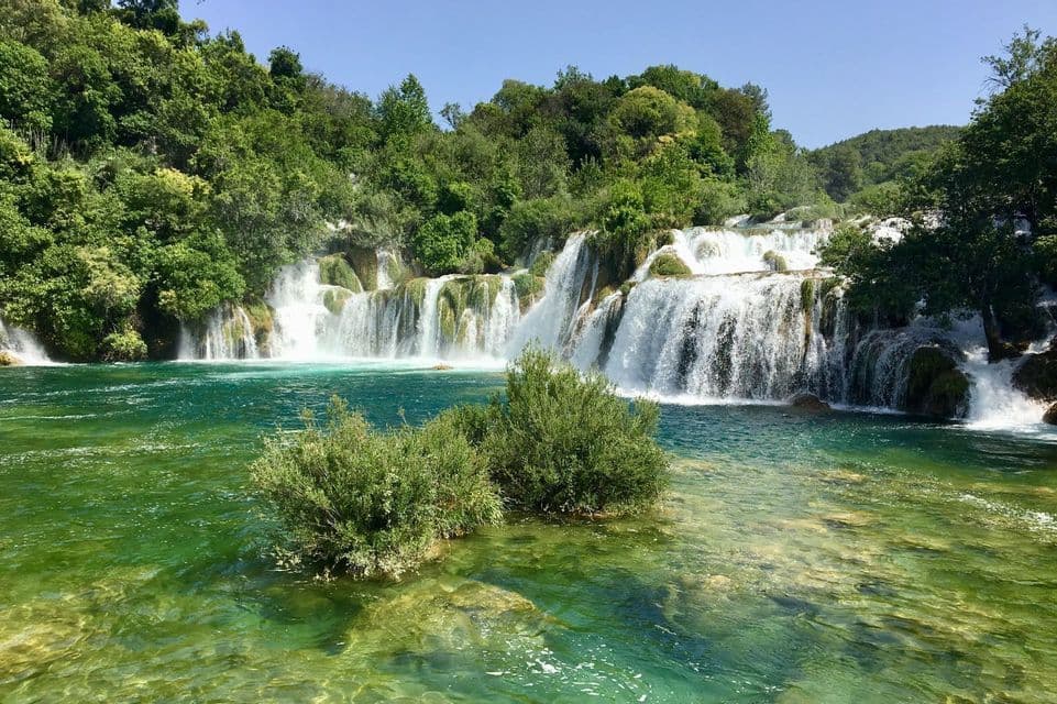 Ein mehrstufiger Wasserfall stürzt über Felsen in einen klaren, grünlichen Fluss, umgeben von einem üppig grünen Wald.