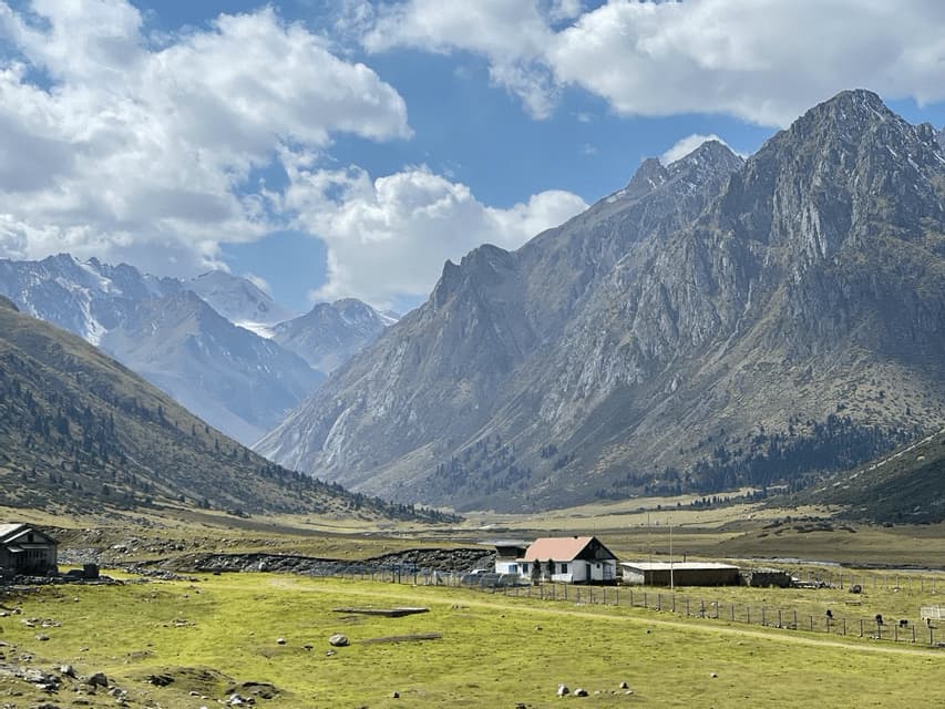 Ein kleines Haus mit rotem Dach liegt in einem weiten grünen Tal am Fuße hoch aufragender, felsiger Berge unter einem bewölkten blauen Himmel.